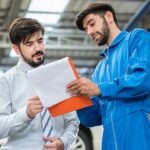 automotive maintenance mechanic young man explain car condition to male customer in garage at auto repair shop, technician pointing needed on vehicle checklist document, after service concept